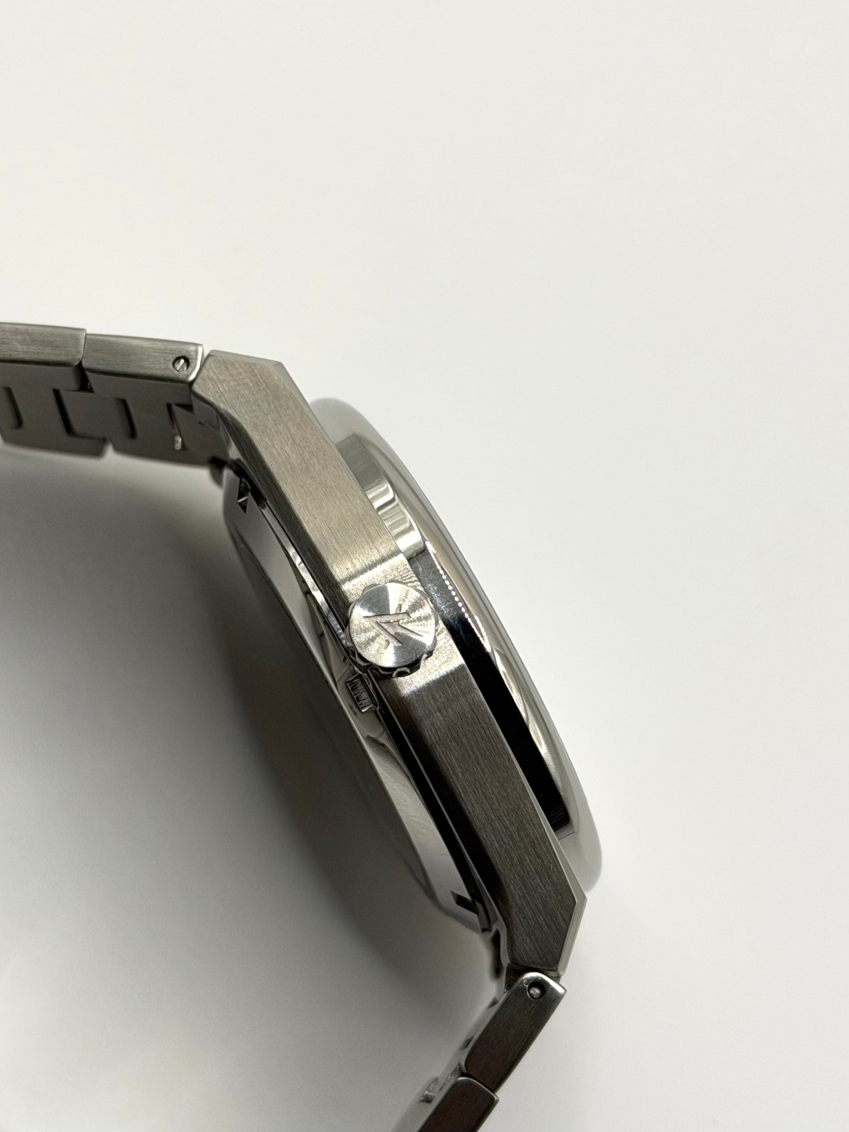 Close-up side profile of a stainless steel watch showing engraved crown with a matching metal link bracelet on a plain light background.